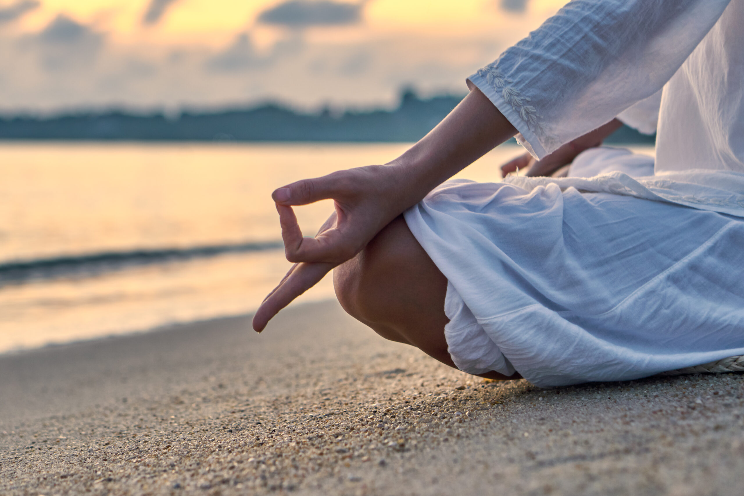 Woman in white clothes and a turban sits on a sandy beach at dawn and practices yoga